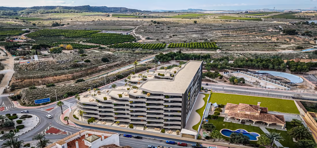 Luchtimpressie van de omgeving (CGI): het gebouw naast de rotonde aan de rand van het stadje, met boomgaarden en heuvels eromheen.